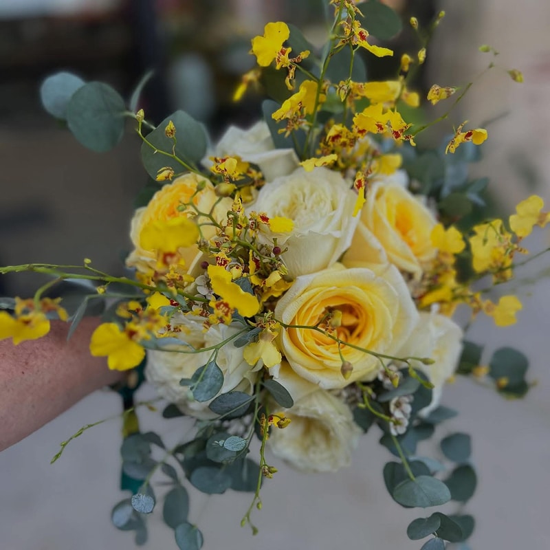 Bouquet Jaune et Blanc: Roses, Orchidées, Eucalyptus Bouquet de mariée jaune et blanc tenu par une main, composé de roses pâles, d'orchidées Oncidium jaunes tachetées et de feuillage d'eucalyptus bleuté.