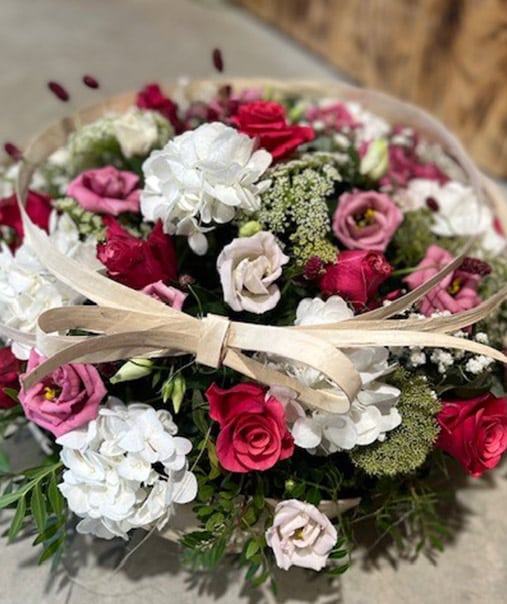 Composition florale ronde et dense, dominée par des roses rouges et blanches, des hortensias blancs, et un ruban beige noué au centre, bouquet élégant et luxuriant.