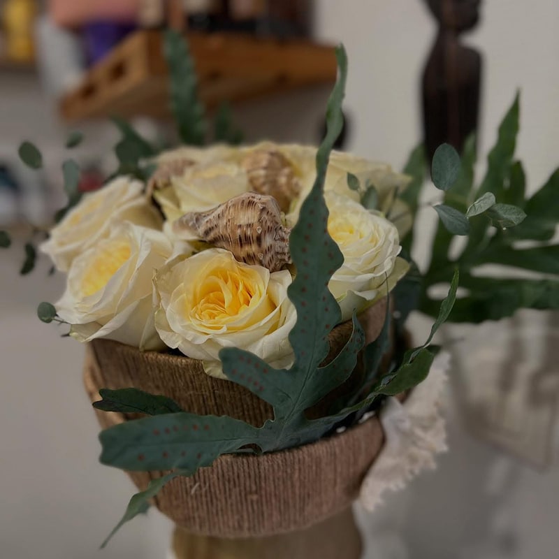 Roses jaunes et coquillages : bouquet marin élégant Composition florale de roses jaunes et crème décorée de coquillages et de feuilles de fougère, dans un contenant texturé en ficelle de jute.
