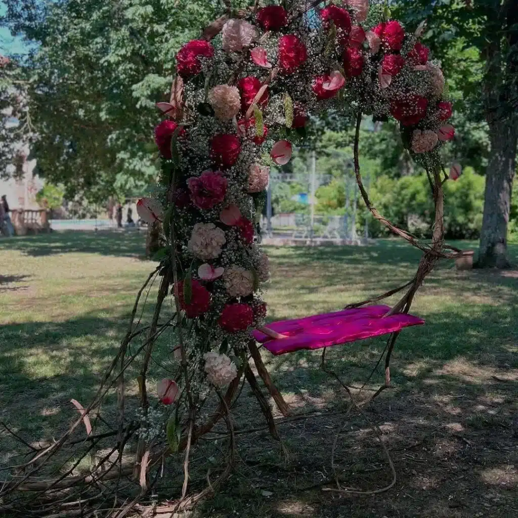 Balançoire florale rouge et rose pour décoration de mariage Balançoire rustique en branches tressées, décorée d'un arc floral rouge, rose et crème. Le siège est agrémenté d'un coussin magenta vif dans un jardin.