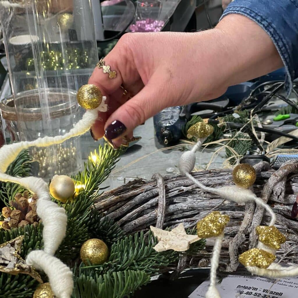 DIY Couronne de Noël : Atelier décoration rustique et doré. Main fixant une boule dorée scintillante sur une couronne de Noël rustique. Décorations DIY, sapin, lumières. Atelier Virazeil.